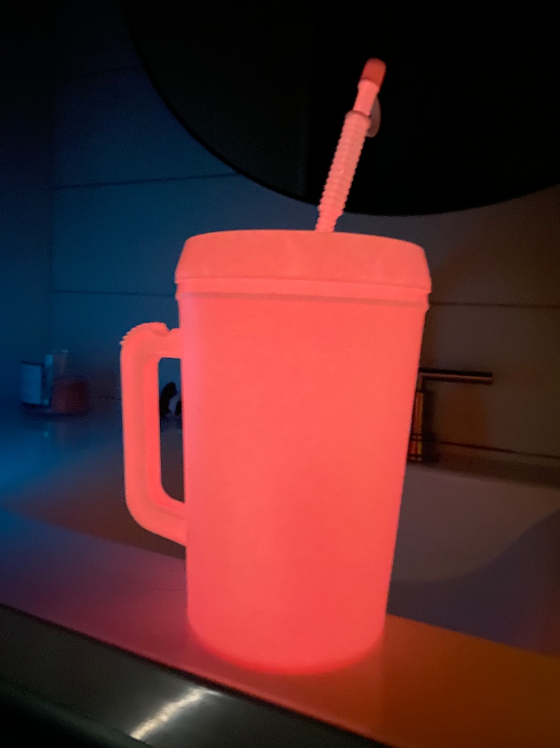 Op de afbeelding: Een witte plastic beker met een handvat en een rietje, die roze gloeit in het donker.