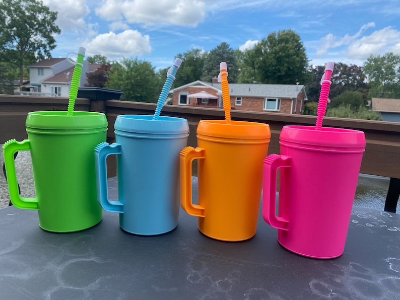 Op de afbeelding: Vier plastic bekers met handvatten en rietjes. De bekers zijn groen, blauw, oranje en roze.