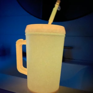 Op de afbeelding: Een gele plastic beker met een handvat en een rietje. De beker staat op een aanrecht.