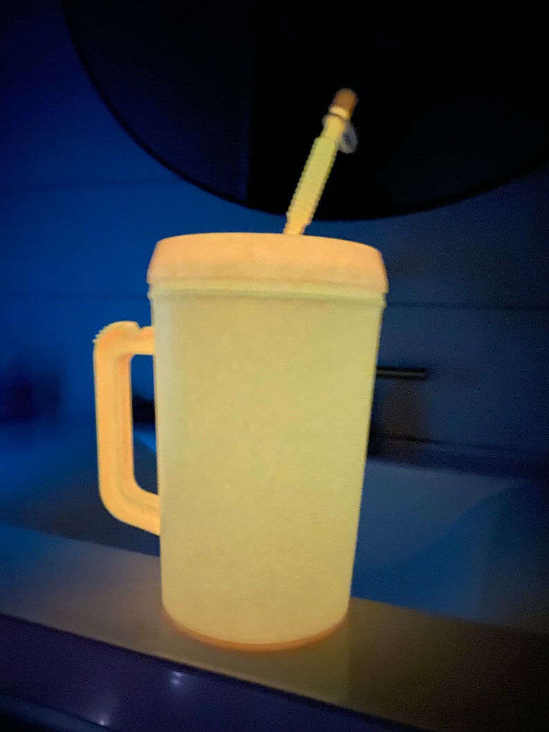 Op de afbeelding: Een gele plastic beker met een handvat en een rietje. De beker staat op een aanrecht.