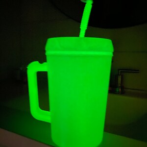 Op de afbeelding: Een groene, in het donker lichtgevende plastic reistumbler met een handvat en een rietje. De tumbler staat op een aanrecht.