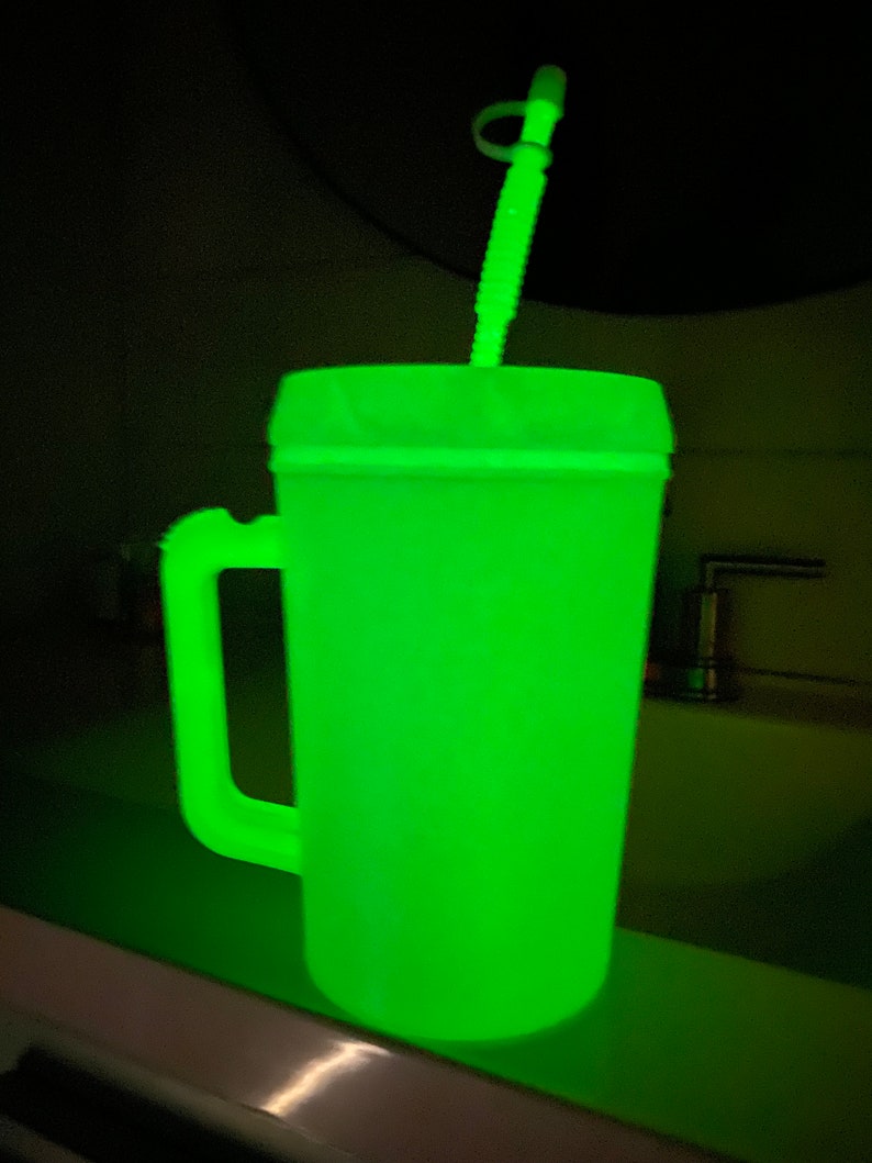 Op de afbeelding: Een groene, in het donker lichtgevende plastic reistumbler met een handvat en een rietje. De tumbler staat op een aanrecht.