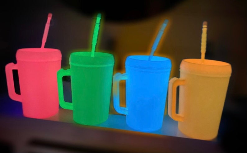 Op de afbeelding: Vier glow-in-the-dark plastic bekers met handvatten en rietjes. De bekers zijn roze, groen, blauw en geel.