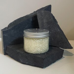 May include: A clear glass jar filled with small white beads, with a silver lid and a small hole in the centre. The jar is sitting on a black rock.