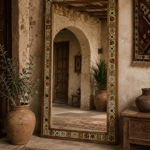 May include: A tall, rectangular floor mirror featuring a thick wooden frame inlaid with a repeating pattern of small, square, multi-coloured tiles in shades of green, yellow, and white. The mirror surface reflects an arched stone doorway.