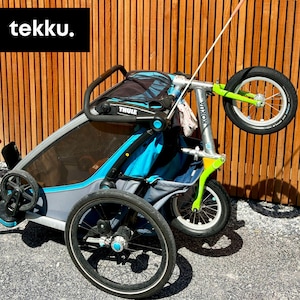 以下が含まれることがあります： 青とグレーのThuleチャイルドキャリア。3つの車輪、黒いハンドル、黒いメッシュフロントが付いています。キャリアには、緑とシルバーのKokua自転車アタッチメントが付いています。「tekku」という単語が左上の黒いボックスにあります。