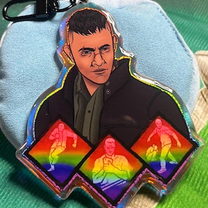 May include: A clear, iridescent keychain featuring a stylized illustration of a person in a black jacket and green shirt. Below are three rainbow-colored diamond shapes with figures in motion. The keychain has a black clasp.