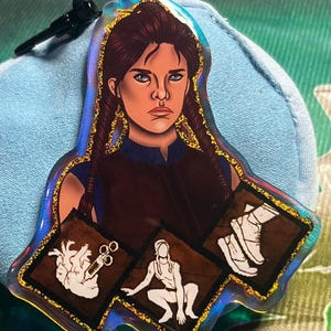 May include: A glittery iridescent keychain featuring a cartoon illustration of a woman with brown hair and a purple shirt. The keychain has three icons: a heart with a syringe, a crouching figure, and a pair of boots.