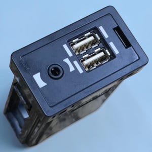 Puede incluir: Un módulo de interfaz electrónica rectangular negro con dos puertos USB apilados y un conector de audio auxiliar circular. La placa frontal incluye iconos impresos en blanco que indican las funciones de los puertos en una carcasa de plástico.