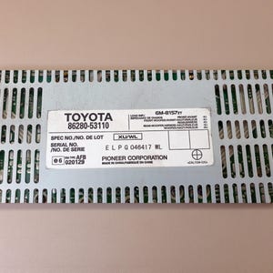 Puede incluir: Un componente amplificador electrónico de metal plateado con carcasa perforada y etiqueta blanca. La etiqueta muestra TOYOTA 86280-53110, GM-8157ZT, PIONEER CORPORATION, el número de serie EL P G 046417 WL y especificaciones técnicas de impedancia.