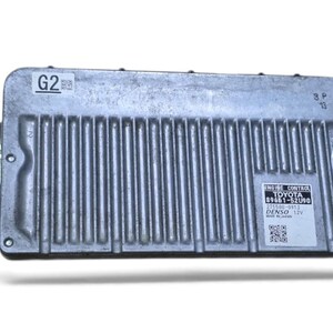 Peut inclure: Un boîtier de commande moteur en métal argenté avec un boîtier rectangulaire nervuré. Il comporte un support de montage de chaque côté et une étiquette blanche affichant le texte TOYOTA 89661-52U90, DENSO 12V et un code QR. Le coin supérieur gauche porte la mention G2.