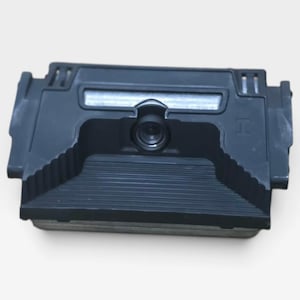 May include: A black automotive backup camera module featuring a central circular lens, a rectangular housing with ribbed plastic detailing, and a metallic mounting bracket visible at the base, designed for vehicle integration.