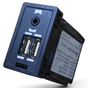 Puede incluir: Un módulo de interfaz electrónica rectangular negro que cuenta con dos puertos USB y un conector de audio auxiliar en el panel frontal. El lateral de la unidad muestra una etiqueta blanca con especificaciones técnicas, incluyendo el texto USB MINI JACK BOX.