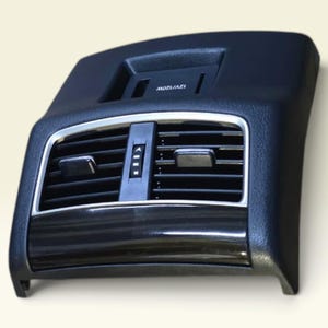 Puede incluir: Un conjunto de ventilación trasera para consola central de automóvil en color negro, con lamas horizontales, un dial de ajuste central y un puerto de alimentación empotrado con la etiqueta 12V/120W, fabricado en plástico texturizado.