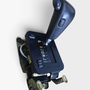 Op de afbeelding: Een zwarte automatische versnellingspook met een getextureerde handgreep en een overdrive-knop. Het paneel toont P, R, N, D, 2, L met gekleurde tekst, een shift lock override-knop en een keuzeschakelaar voor de standen PWR, ECT en NORM.