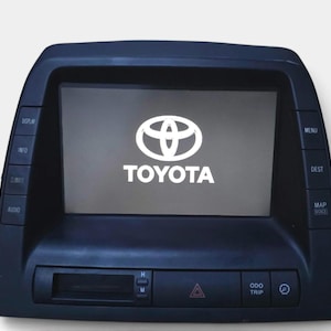 May include: A black Toyota automotive navigation and infotainment display unit featuring a central screen with the Toyota logo. The unit includes side buttons labelled Display, Info, Climate, Audio, Menu, Dest, and Map Voice, plus bottom controls.