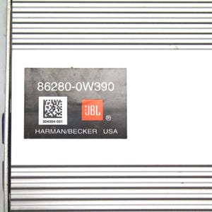 Puede incluir: Una carcasa de amplificador electrónico de metal plateado con una superficie acanalada. Una etiqueta rectangular negra está adherida al centro, mostrando el texto 86280-0W390, un código QR, el logotipo de JBL y el texto HARMAN/BECKER USA.