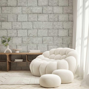 Może przedstawiać: Białe, pluszowe, duże krzesło o okrągłym, puszystym kształcie. Krzesło stoi na jasnobrązowym dywanie przed białą ceglaną ścianą. Przed krzesłem znajduje się mały, okrągły podnóżek.