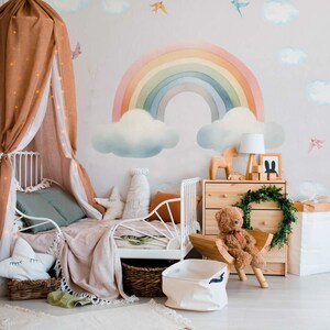 May include: A white metal bed frame with a brown and white blanket and a rainbow wall decal with clouds. The bed is decorated with pillows and a stuffed animal.