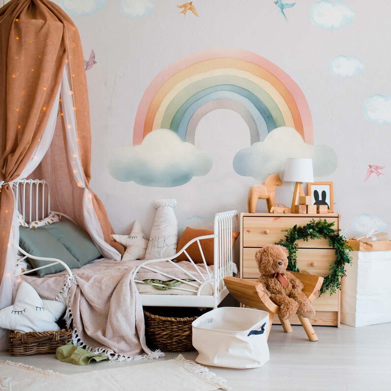 May include: A white metal bed frame with a brown and white blanket and a rainbow wall decal with clouds. The bed is decorated with pillows and a stuffed animal.