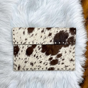 Könnte beinhalten: Eine rechteckige Clutch mit einem braun-weißen Kuhfellmuster. Die Kanten der Tasche sind mit einer Reihe kleiner, silberfarbener Metallnieten verziert, die den Rand der Klappe und den unteren Teil des Hauptkörpers der Tasche säumen.