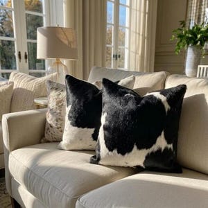 May include: Two square throw pillows featuring a black and white cowhide pattern with a textured fur finish. The pillows are arranged on a light beige upholstered sofa, positioned next to a third pillow with a speckled brown and white pattern.