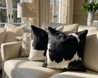 May include: Two square scatter cushions featuring a black and white cowhide pattern with a textured fur finish. The cushions are arranged on a light beige upholstered sofa, positioned next to a third cushion with a speckled brown and white pattern.