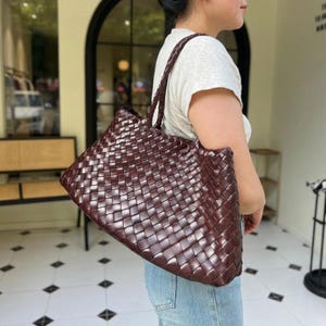 Può includere: Una grande borsa tote color bordeaux intenso caratterizzata da un intricato motivo in pelle intrecciata su tutto l'esterno. La borsa presenta un unico spallaccio spesso e intrecciato e una silhouette rettangolare spaziosa con un bordo superiore strutturato e ripiegato.