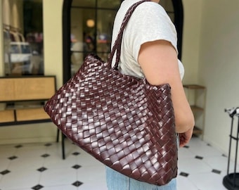 May include: A large, deep burgundy tote bag featuring an intricate woven leather pattern throughout the exterior. The bag has a single, thick braided shoulder strap and a spacious, rectangular silhouette with a structured, folded top edge.