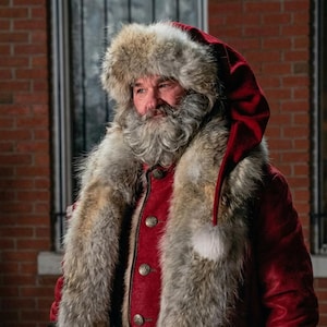 May include: A person with a full gray beard wearing a red velvet coat and a matching red hat with a long tail. The coat and hat are trimmed with thick, textured gray and white fur. The coat features round, metallic buttons down the front.