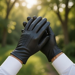 May include: A pair of black leather gloves, with visible stitching details, are held together against a blurred green background. The gloves are worn over a white dress shirt, creating a classic and elegant look. The gloves appear to be new.