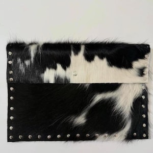 May include: A rectangular clutch made of cowhide with a black and white spotted pattern. The top flap and bottom edge are adorned with silver-colored studs. The bag has a natural, textured appearance.