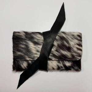 May include: A rectangular clutch with a black leather strap closure. The clutch is made of cowhide with a black and white spotted pattern. The strap wraps around the clutch and is secured with a knot.