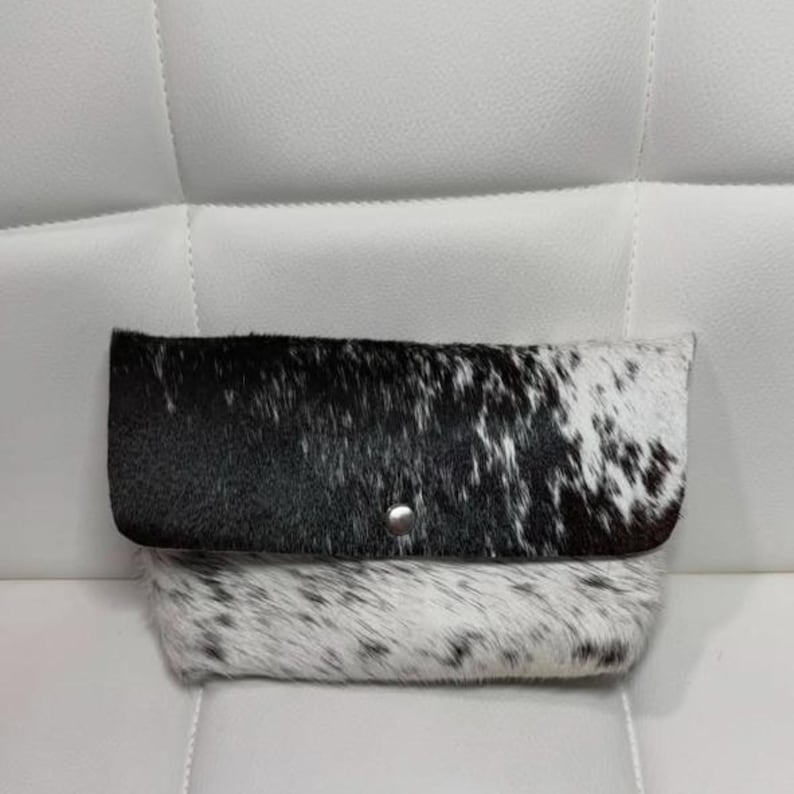 May include: A black and white cowhide clutch with a fold-over flap secured by a silver snap. The bag features a natural spotted pattern, with a mix of black and white fur. The clutch is rectangular and appears to be made of genuine leather.