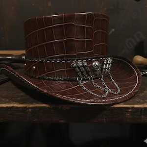 Puede incluir: Un sombrero de copa de cuero marrón con textura de piel de cocodrilo. El sombrero presenta una banda decorativa con dijes plateados de calaveras y balas, y una cadena plateada. El ala está cosida con hilo blanco.