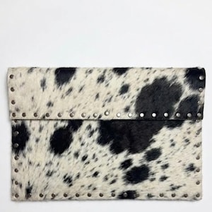 May include: A rectangular clutch with a cowhide pattern in black and white. The edges are lined with small, silver-colored rivets. The flap is folded over and secured, creating a stylish accessory.