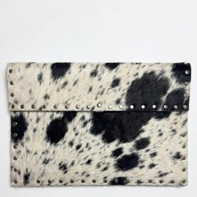 May include: A rectangular clutch with a cowhide pattern in black and white. The edges are lined with small, silver-colored rivets. The flap is folded over and secured, creating a stylish accessory.