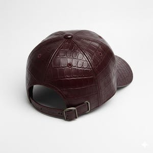 May include: A burgundy baseball cap with a textured, crocodile-like pattern. The cap features a curved brim, a button on top, and an adjustable strap with a metal buckle. The hat is made of a shiny material.
