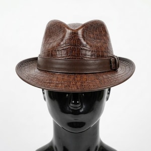 May include: A brown fedora hat featuring a textured crocodile skin pattern across the crown and brim. The hat includes a smooth, solid dark brown leather band encircling the base of the crown, finished with a small rectangular buckle detail.