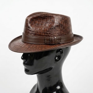 May include: A brown fedora hat featuring a textured crocodile skin pattern across the crown and brim. The hat includes a smooth, solid brown leather band encircling the base, complete with a small rectangular buckle detail on the side.
