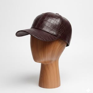 May include: A burgundy baseball cap with a textured, crocodile-like pattern. The cap is displayed on a wooden mannequin head. The hat has a curved brim and a structured crown, offering a stylish accessory.