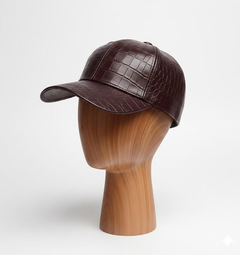 May include: A burgundy baseball cap with a textured, crocodile-like pattern. The cap is displayed on a wooden mannequin head. The hat has a curved brim and a structured crown, offering a stylish accessory.