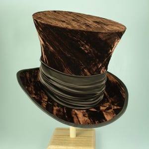 Pode incluir: Um chapéu de copa de veludo castanho com uma faixa e aba pretas. O chapéu é exibido em um suporte de madeira sobre um fundo azul claro. A textura e a forma do chapéu sugerem um estilo vintage ou teatral.