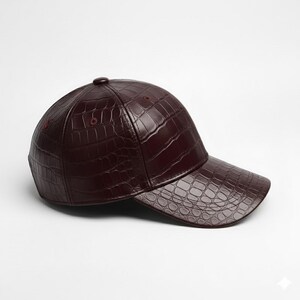 May include: A burgundy baseball cap with a textured, crocodile-like pattern. The cap features a curved brim and a button on top. The hat is made of a shiny material.