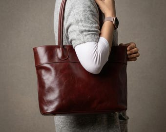 May include: A deep burgundy leather tote bag with a smooth, polished finish. The bag features two long, slender shoulder straps attached to a horizontal stitched panel across the top. The design includes a wide main compartment and gold-tone hardware.