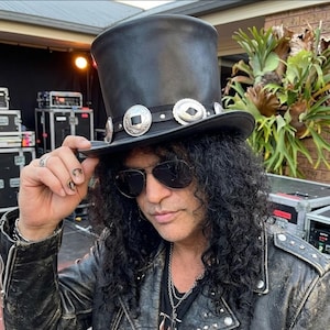 May include: A black leather top hat featuring a black band adorned with circular silver-toned metal concho accents. The hat has a tall, cylindrical crown and a wide, slightly curved brim, worn with dark-tinted sunglasses and a black leather jacket.