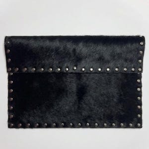 May include: Black clutch made of textured, animal-hair material. The rectangular bag features a flap closure and is embellished with silver-colored studs along the edges. The bag is a stylish accessory.