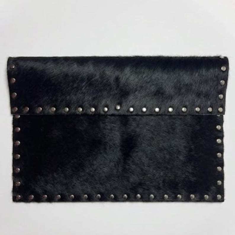 May include: Black clutch made of textured, animal-hair material. The rectangular bag features a flap closure and is embellished with silver-colored studs along the edges. The bag is a stylish accessory.
