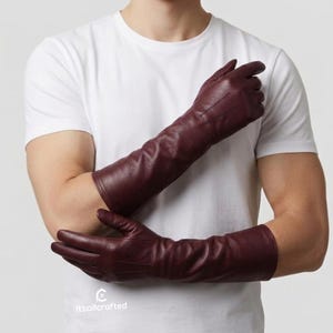 May include: A pair of long, burgundy leather gloves. The gloves extend past the elbows and have a smooth texture. The gloves are shown against a white t-shirt.