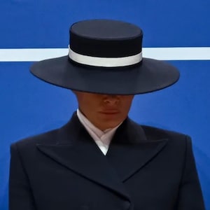 Può includere: Un cappello nero con tesa piatta e fascia bianca, indossato con un abito scuro e camicia bianca. Il cappello ha una corona cilindrica. Lo sfondo è blu con una striscia orizzontale bianca.